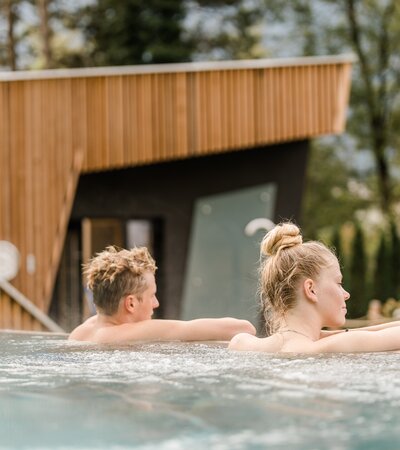 Wellness im Val Blu in Bludenz | © Hefti Fotografie Zwei Personen, ein Mann und eine Frau, entspannen im Outdoor-Pool des Wellnessbereichs im Val Blu; im Hintergrund Garten, Bäume und eine Sauna bei Tageslicht. | © Hefti Fotografie
