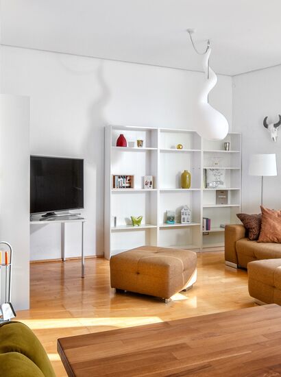Wohnbereich einer Ferienwohnung mit Couch, Fernseher, dekoriertem Bücherregal, Schwedenofen und Designerlampe; der Esstisch mit Stühlen ist am Bildrand angeschnitten sichtbar. | © Casa Allegria