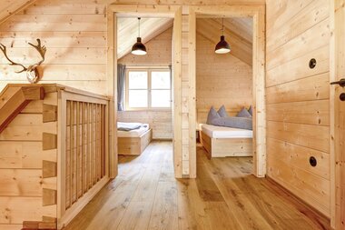Blick vom oberen Flur eines Chalets durch zwei Türen in zwei schlicht und modern eingerichtete Schlafzimmer; Innenräume vollständig aus Holz mit warmem Licht und minimalistischer Ausstattung. | © Casalpin.com