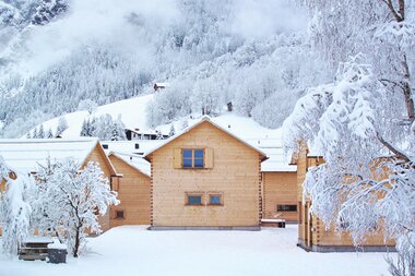 Verschneite Winterlandschaft mit mehreren Holzchalets im Vordergrund; dahinter bewaldete, schneebedeckte Berge – ruhige, alpine Szenerie. | © Casalpin.com