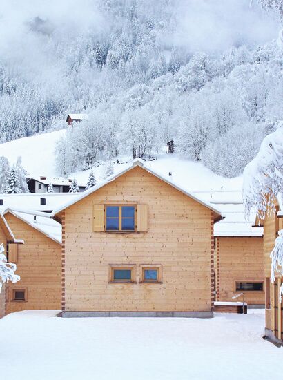 Verschneite Winterlandschaft mit mehreren Holzchalets im Vordergrund; dahinter bewaldete, schneebedeckte Berge – ruhige, alpine Szenerie. | © Casalpin.com