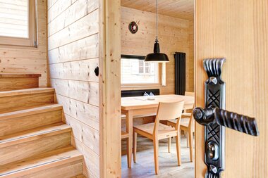 Innenaufnahme einer gemütlichen Ferienwohnung mit viel Holz; Blick aus dem Vorraum auf eine hölzerne Treppe ins Obergeschoss und durch die leicht geöffnete Tür in das Esszimmer. | © Casalpin.com