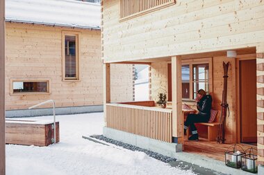 Ein Holzchalet in verschneiter Winterlandschaft; eine Frau sitzt auf der Veranda und trinkt eine Tasse Tee. | © Casalpin.com